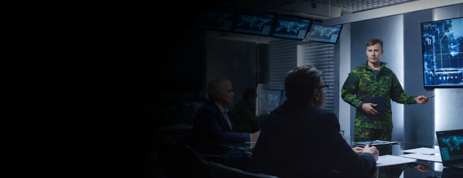 A man in army fatigues at the front of a classroom, gesturing to information on a Secure III LCD panel.