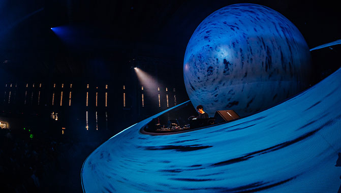 A side view of a DJ situated in a booth located inside a projection mapped replica of the planet Saturn. 