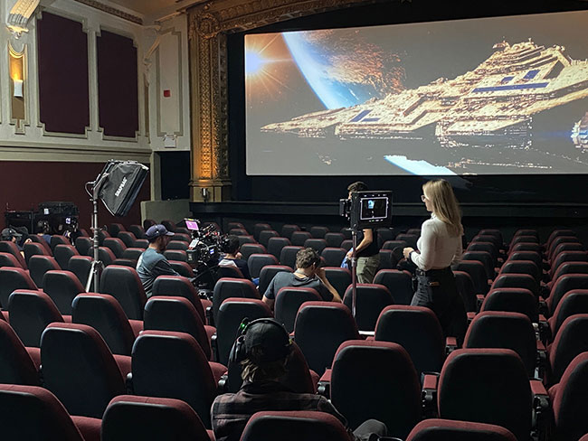 Five people are sitting and standing in a brightly lit movie theatre. There is a futuristic looking spaceship shown on the screen.
