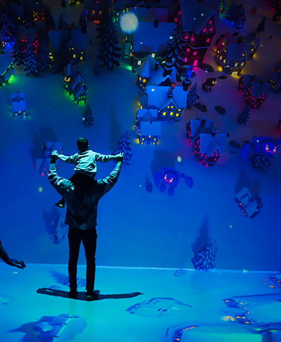 A person with a child on their shoulders looks at a nighttime scene of houses covered in snow that is projection mapped onto a wall and floor.