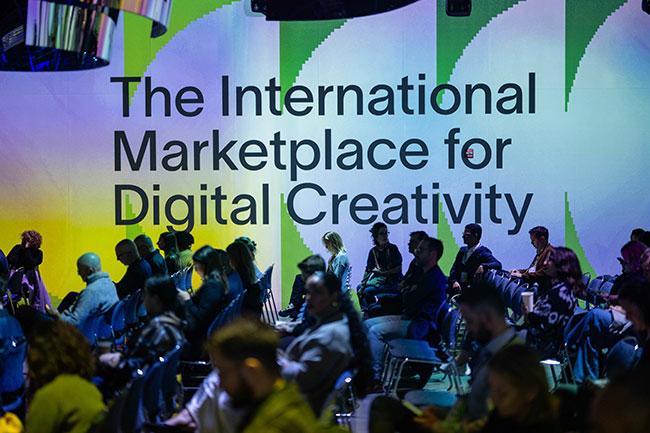 People sitting in chairs in a large room with the text &ldquo;The International Marketplace for Digital Creativity&rdquo; projected onto the wall.