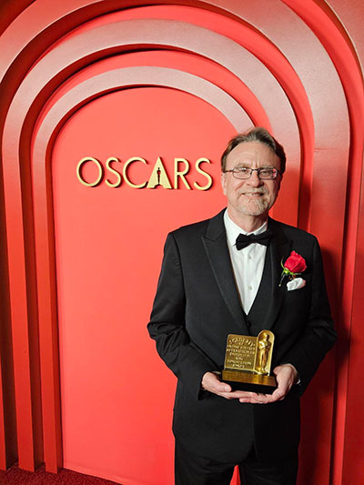 Man dressed in a tuxedo holding a gold award standing in front of a red wall with the word &ldquo;Oscars&rdquo; on it.