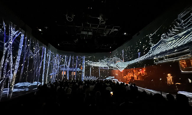 A tree and snow-filled landscape is projected onto three walls of a room while a group of people look on.