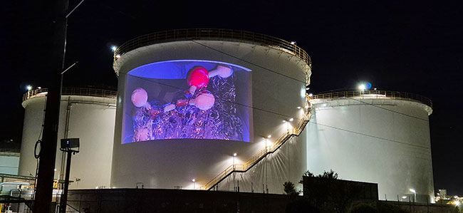 A large fuel storage tank is lit by a single projector at night.