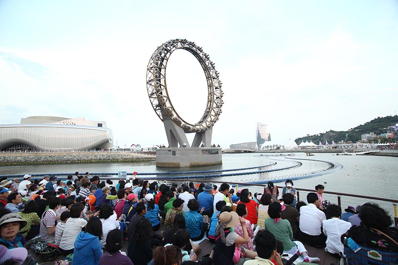 People sit on the ground in front of a pond with a large circular structure in the center.