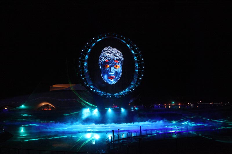 A large blue face is projected in the middle of a ring of water in the middle of a pond.