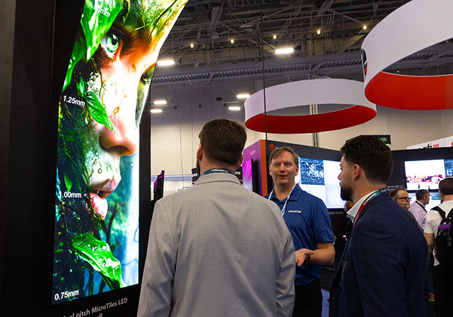 Three people stand in front of a vertical MicroTiles image display the the different pixel pitches overlayed on the image.