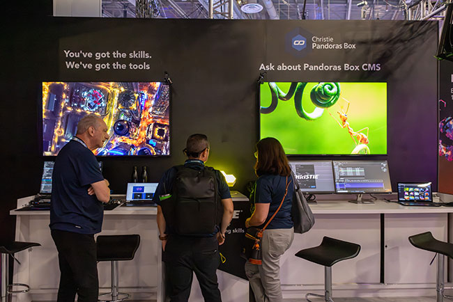 Three people stand in front of a desk with monitors in a trade show booth.