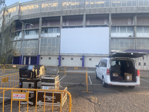 A movie screen is mounted to the side of a building with a projector and minivan in front of it.