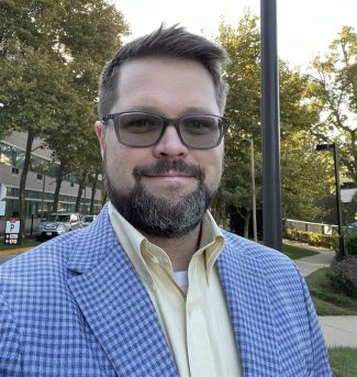  close-up of a man standing outside wearing sunglasses and a blue suit jacket.