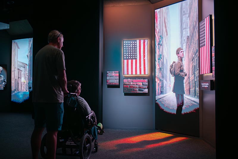 A person standing and a person in a wheelchair look at the image of a man shown on an LED display.